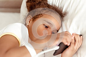 Child, technology and internet concept - little girl lying in bed with smartphone at home