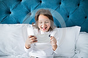 Child, technology and internet concept - little girl lying in bed with smartphone playing games at home