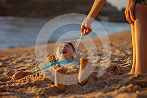 Child, tickling sibling on the beach on the feet with feather, kid cover in sand, smiling, laughing