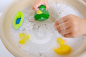 child, toddler 3 years old plays with rubber green, yellow ducks for swimming, child's toy in soapy foam, close-up of hand,