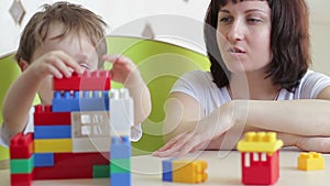The child, together with her mother, builds a house of colored Lego blocks at the table. Child development.