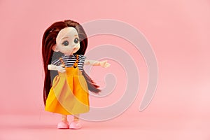 child toy doll with dark hair in an orange dress standing on a pink background. Plastic children's toy. a plastic doll indicates