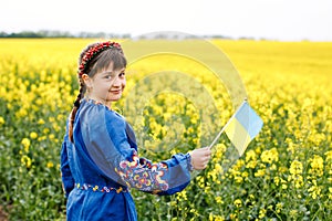 Child with Ukrainian flag in rapeseed field. A girl in an embroidered shirt runs across the field with the Ukrainian flag in her