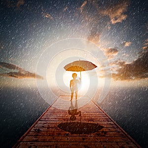 Child with umbrella standing alone wooden jetty in rain looking at the sea.