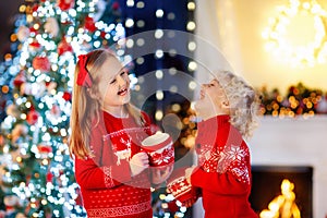 Child under Christmas tree at home. Little boy and girl in knitted sweater with Xmas ornament drink hot chocolate. Family with