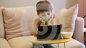 Child using tablet pc on bed at home. Cute boy on sofa is watching cartoon, playing games and learning from laptop. Education, fun