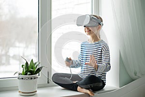 A child with a virtual reality headset is sitting at the table indoors at home. A girl exploring the world of virtual reality