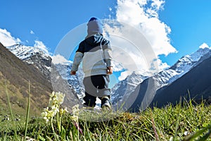 A child walks against the background of snowy mountains, in the foreground flowers of the field