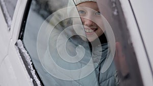 A child in warm winter clothes looks and smiles at the camera through the car window. Winter portrait of a little girl