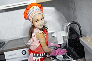 Child washes laptop in sink on kitchen. Wearing red apron, cook hat and pink gloves.