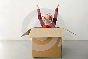 A child wearing funny Christmas glasses and Santa hat raised her hands up while sitting in a cardboard box against white