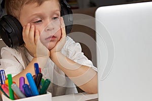 A child wearing headphones looks wearily at the laptop screen.