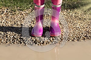 Child wearing new pink rain rubber boots standing near muddy puddle