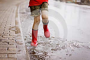 Child wearing red rain boots jumping into a puddle. Close up. Kid having fun with splashing with water. Warm heavy