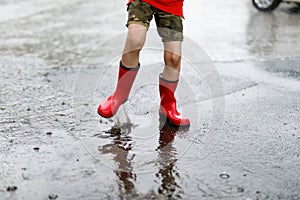 Child wearing red rain boots jumping into a puddle. Close up. Kid having fun with splashing with water. Warm heavy