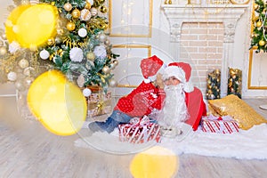 A child whispers to Santa Claus in the ear near the Christmas tree in the yellow and white Christmas interior, a happy child talks