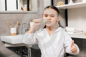 child in white bathrobe brushing teeth and showing thumb up photo