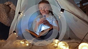 child in a wigwam tent play at night with a flashlight and read a book. happy family kid dream concept. child reading