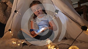 child in a wigwam tent play at night with a flashlight and read a book. happy family kid dream concept. child reading