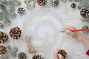 Child writes the letter to Santa Claus. Child`s hands, the sheet of paper, red pencil and Christmas decorations pine