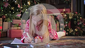 Child writes letter with wishes for gift for Santa Claus. Small blond girl in red sweater lies on floor near Christmas