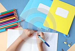 A child writes with a pen in a student`s notebook on a table with writing materials, top view