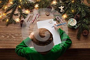 Child Writing Christmas Letter to Santa Claus. Top View of Kid sitting at Wooden Table with Xmas Tree Decoration Lights and Empty