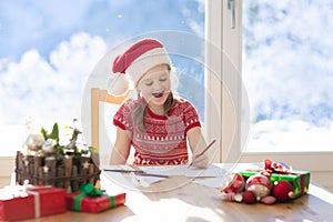 Child writing letter to Santa on Christmas eve. Kids write Xmas present wish list. Little girl sitting in decorated living room