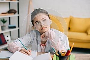 Child writing in notebook and looking away while doing schoolwork at home