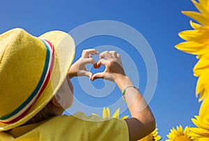 Child in a yellow hat against the background of the sky and blooming sunflowers holds hands in the shape of a heart