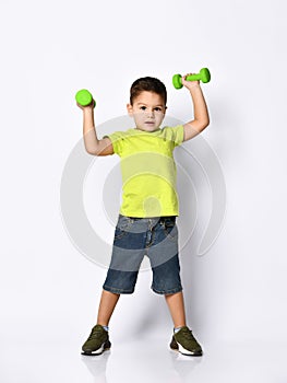 Child in yellow t-shirt, denim shorts, khaki sneakers. Smiling, raised hands up holding green dumbbells, posing isolated on white