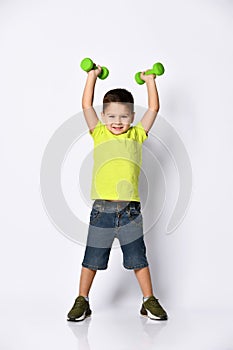 Child in yellow t-shirt, denim shorts, khaki sneakers. Smiling, raised hands up holding green dumbbells, posing isolated on white