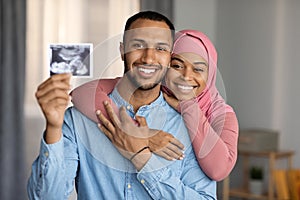 Childbirth concept. Happy black muslim couple expecting baby, showing sonography at camera
