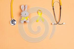 Childhood Cancer Awareness Golden Ribbon with toy bunny and stethoscope on yellow background with copy space.