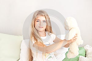 Childhood concept. Little girl with blond long hair playing in bed in a white bedroom. Toddler with a toy. happy girl playing with