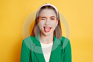 Childish behavior. Playful positive woman wearing green jacket winking to camera showing tongue out posing isolated over yellow