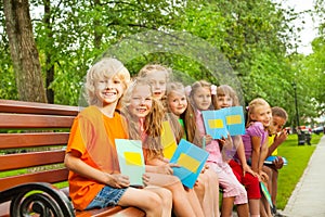 Children with blue notebooks sit in row on bench