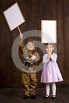 Children boy are dressed as soldier in retro military uniforms and girl in pink dress. They`re holding blank posters for veterans