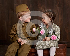 Children boy are dressed as soldier in retro military uniforms and girl in pink dress sitting on old suitcase, dark wood backgroun