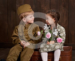 Children boy are dressed as soldier in retro military uniforms and girl in pink dress sitting on old suitcase, dark wood backgroun