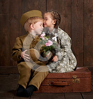 Children boy are dressed as soldier in retro military uniforms and girl in pink dress sitting on old suitcase, dark wood backgroun