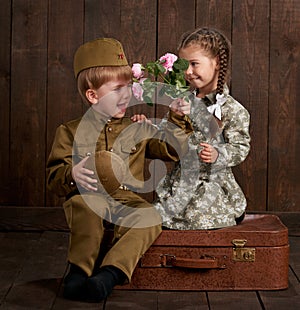 Children boy are dressed as soldier in retro military uniforms and girl in pink dress sitting on old suitcase, dark wood backgroun