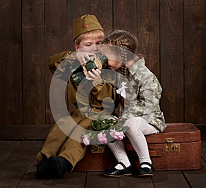Children boy are dressed as soldier in retro military uniforms and girl in pink dress sitting on old suitcase, dark wood backgroun
