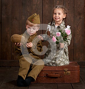 Children boy are dressed as soldier in retro military uniforms and girl in pink dress sitting on old suitcase, dark wood backgroun