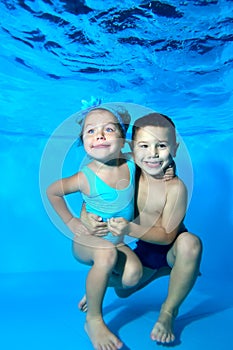 Children, a boy and a girl, smile and pose under the water in the pool on a blue background. Portrait. Close-up