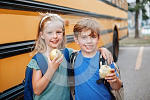 Children boy and girl students friends eating apples healthy snack by yellow school bus outdoor. Education and back to school in