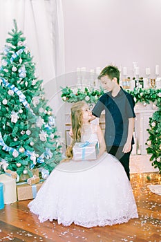 Children, a boy and a girl in a white ball gown near a Christmas tree in holiday dresses with gifts and silver confetti.