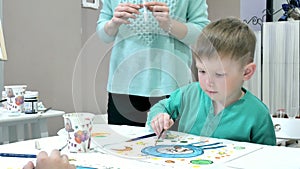 Children boys sitting together around the table in classroom and drawing. With them is their young and beautiful teacher