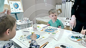 Children boys sitting together around the table in classroom and drawing. With them is their young and beautiful teacher