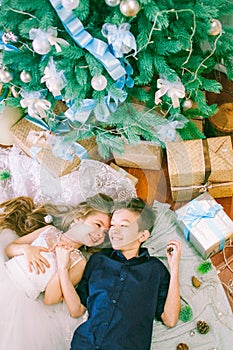 Children, brother and sister, are lying under the Christmas tree in holiday dresses with gifts and silver confetti.